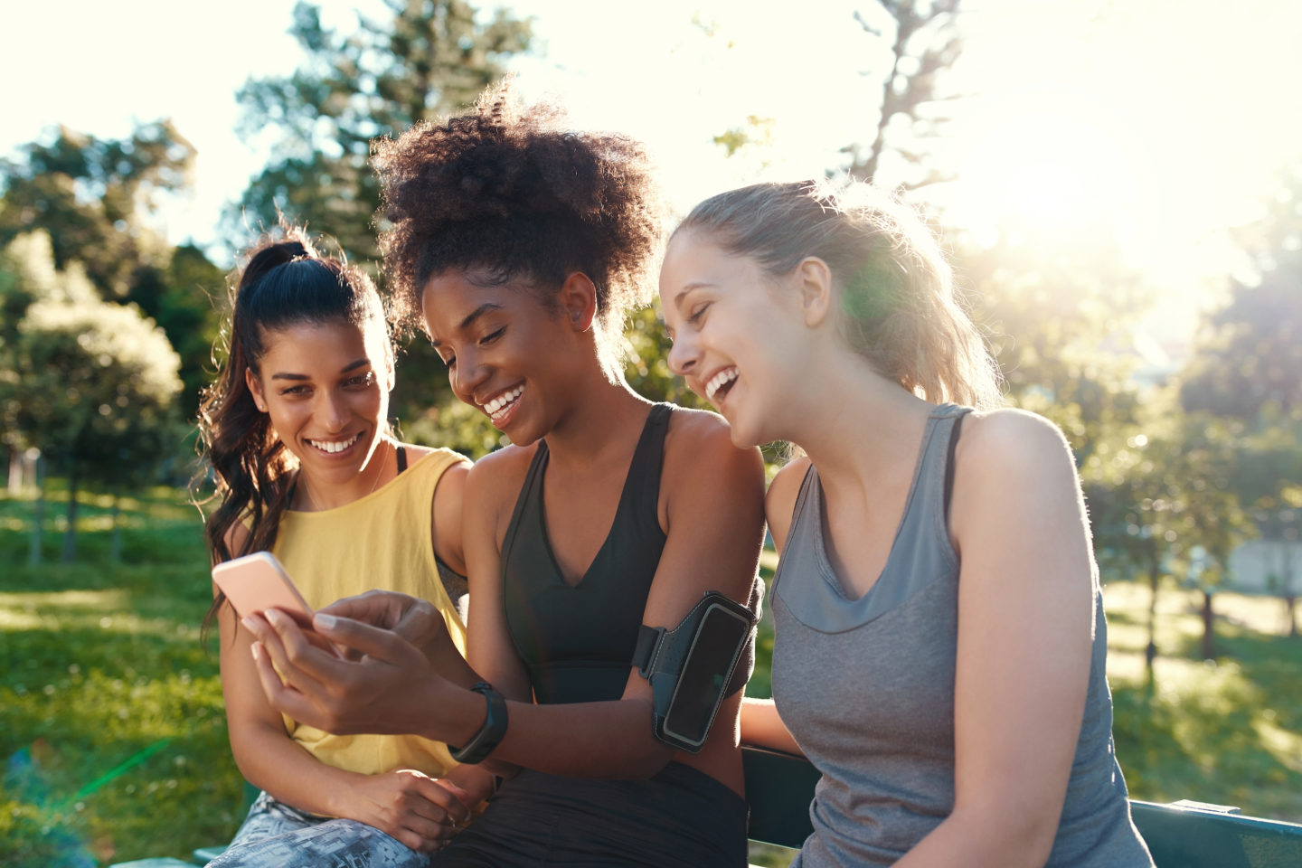 Smiling female diverse friends looking at mobile phone smiling and having fun in the morning at the park - friends laughing together