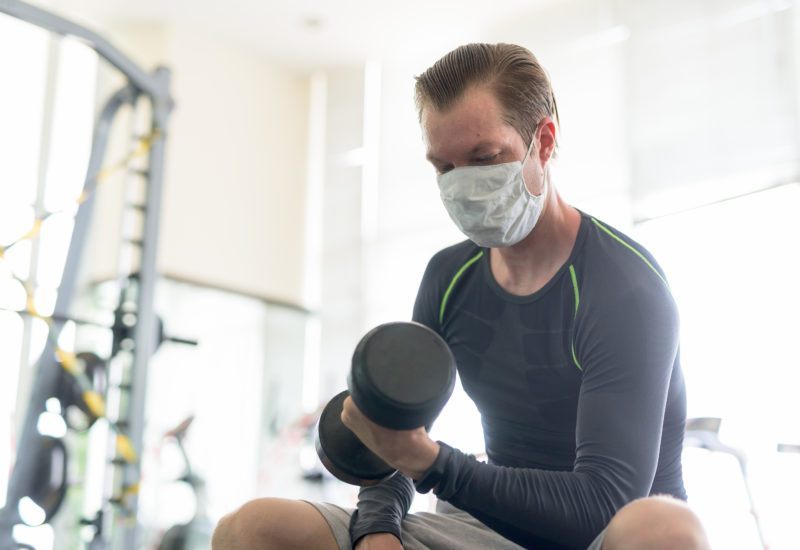 Young man with mask sitting while exercising with dumbbell at gym during corona virus covid-19