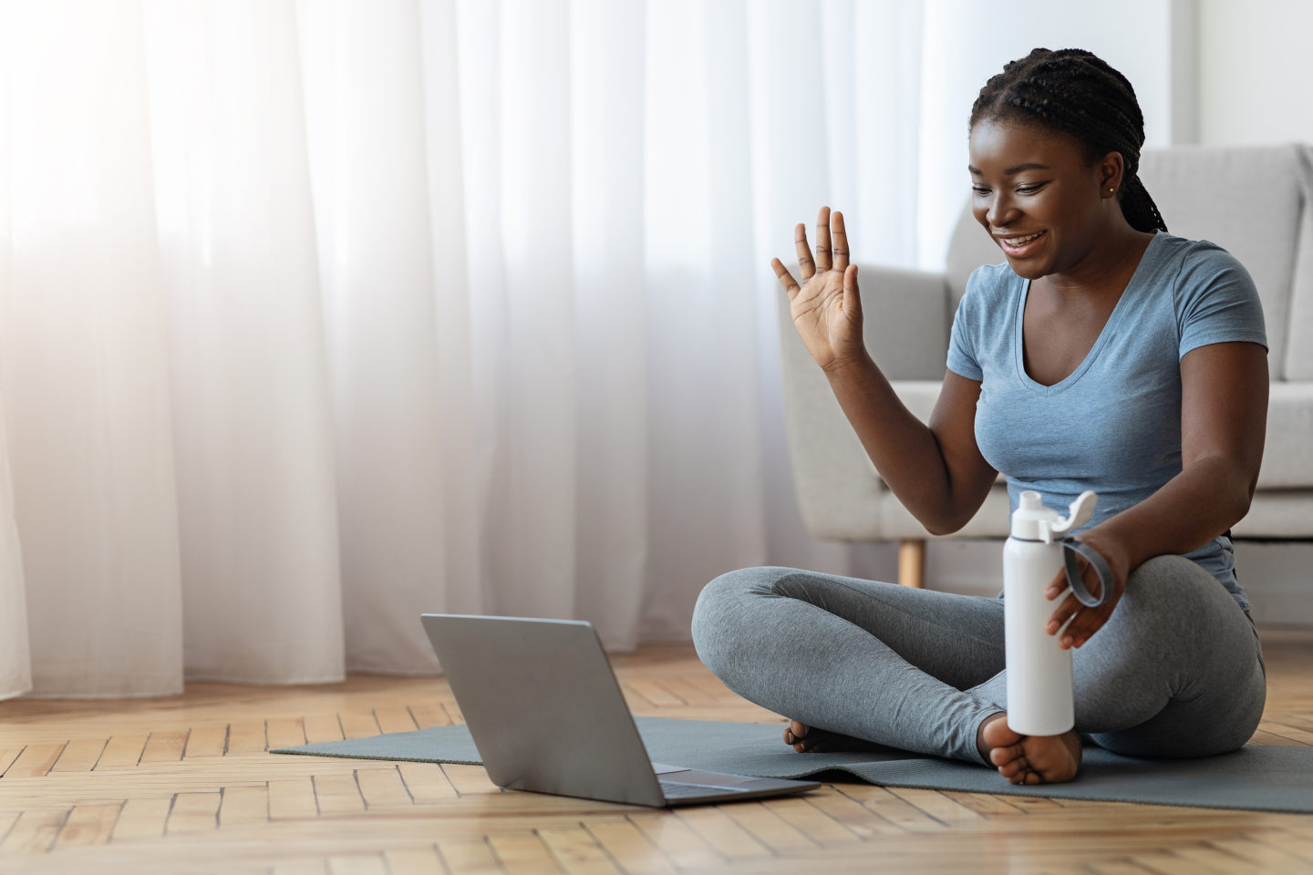 Smiling Black Woman In Sportswear Having Online Fitness Training With Laptop At Home, Waving Hand At Computer Webcam, Greeting Coach, Exercising On Yoga Mat In Living Room, Enjoying Healthy Lifestyle