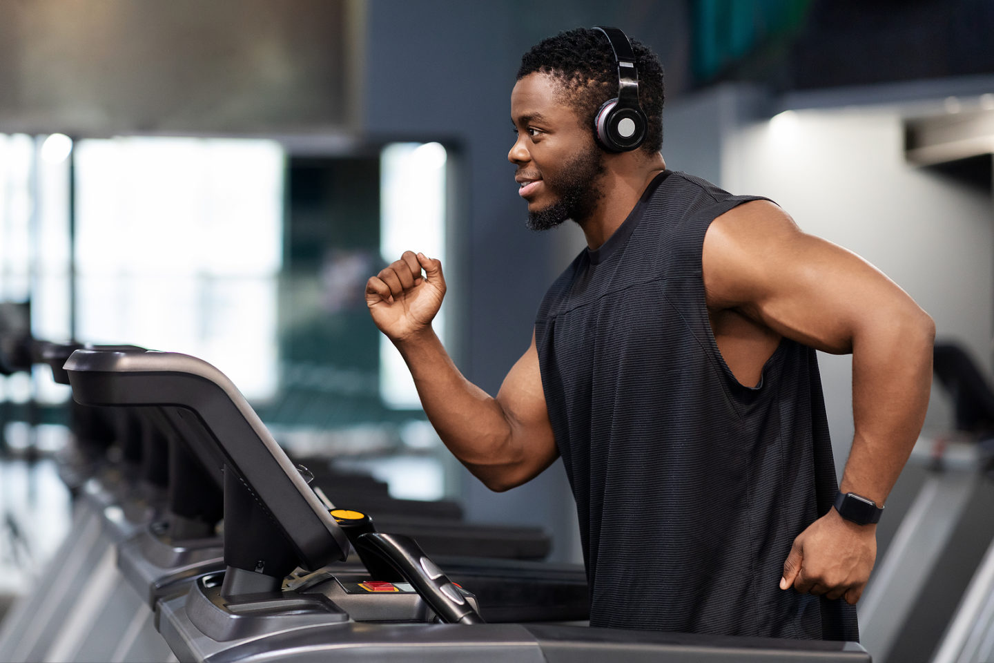 Jogging workout. Joyful black man in modern wireless headphones running on treadmill at gym, looking at free space. Motivated african american guy in black sportswear spending time at gym, jogging
