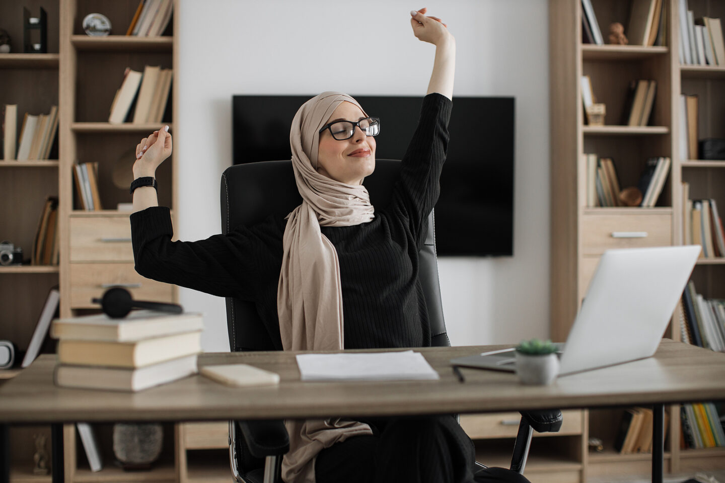 Smiling muslim woman relaxing by stretching her body while sitting in front of pc computer laptop at the wooden working table over comfortable office.