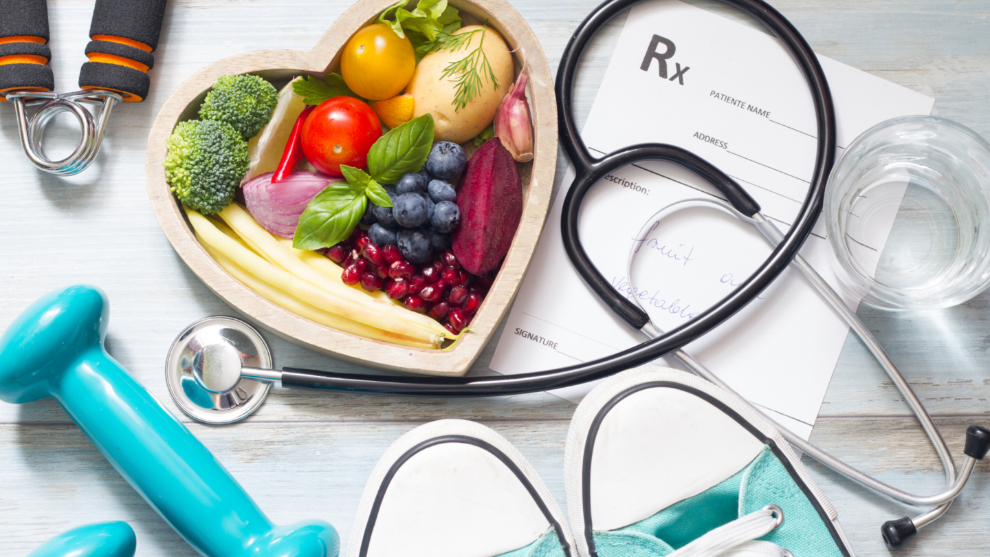 An overhead view of a heart shaped bowl filled with healthy fruits, a set of light blue hand weights, a stethascope all siting on top of Rx medical documents