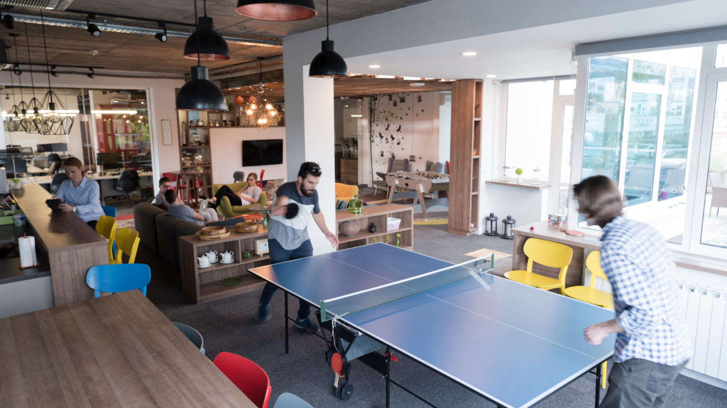 A diverse group of employees engagement in social activities in their workplace such as playing ping pong and socializing in the cafe.